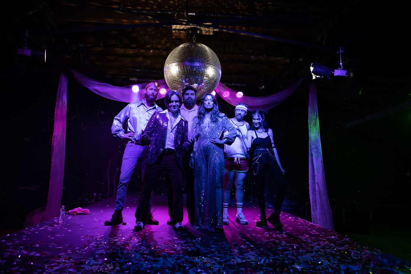 Six actors posing under purple lights and a giant disco ball.